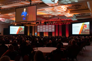 Conference general session featuring people sitting at round tables, four large screens, and speaker on stage.