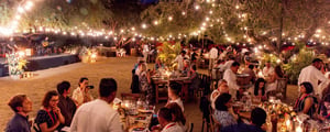 corporate event planner image showing several tables of people enjoying dinner, string lights overhead, and a band playing in the background.