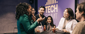 group of people talking around a table at a conference with a sign in the back that says tech summit.