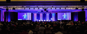 large conference stage with two screens and speaker onstage that has purple lighting and an audience watching